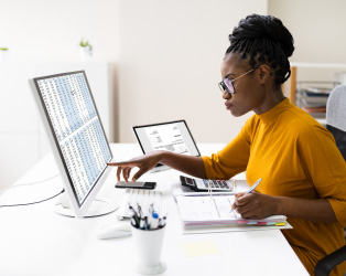 Imagem de uma jovem negra de óculos, sentada em uma mesa de perfil, em um fundo claro, usando uma blusa amarela, trabalhando em dois computadores em planilhas administrativas