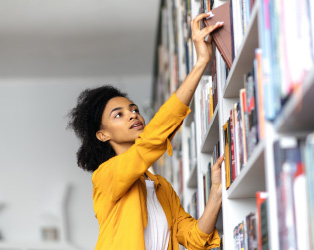 Imagem de uma jovem negra, vestindo uma blusa amarela, em uma biblioteca pegando um livro em uma estante