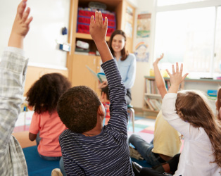 Imagem de uma sala de aula com crianças de costas com as mãos levantadas em frente a professora que está sorrindo de frente para elas.