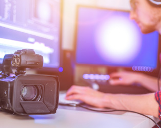 Imagem de uma câmera de filmagem em destaque e ao fundo desfocado uma pessoa trabalhando em uma ilha de edição de vídeo com dois monitores e diversos aparelhos audiovisuais.