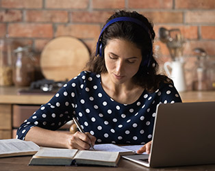 Mulher branca de fone de ouvidos, cabelos castanhos escuros, vestindo uma blusa azul escuro com bolinhas brancas, em uma mesa de estudos com um notebook aberto, escrevendo em um caderno com um fundo desfocado.