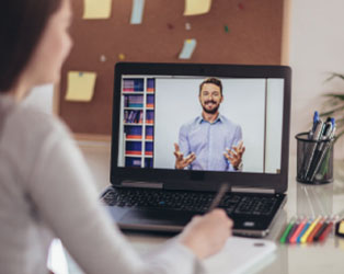 Imagem de uma aluna de costas desfocada, assistindo uma aula em um computador com o professor em tela explicando e ao fundo um quadro com post its desfocados.