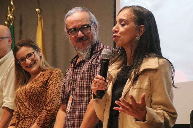 Três pessoas em pé no palco; uma mulher fala ao microfone enquanto as outras a acompanham.