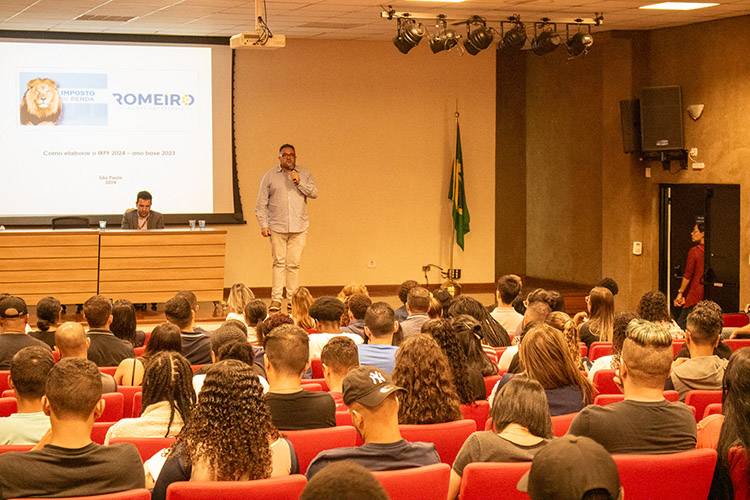 Professor, no palco, de frente para o público, ao microfone, fala com a plateia.