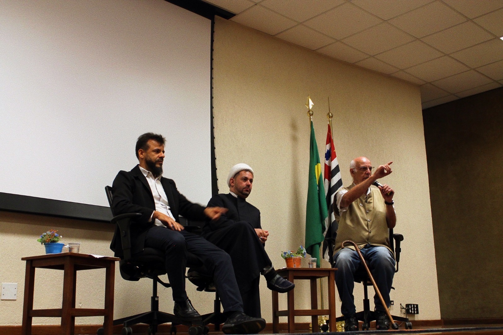Pe. Leomar, Pe. Júlio Lancellotti e o Sheik Rodrigo Jalloul em cima do palco no auditório do UNIFAI. Pe. Júlio fala ao microfone.