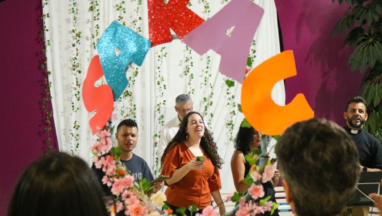 Padre Leomar, ao microfone, acompanhado de aluna, cantam para o público do sarau.