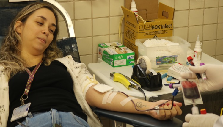 Professora deitada na maca com braço estendido doando sangue.