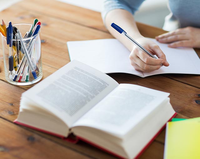 Imagem de uma pessoa escrevendo em um caderno em branco sob uma mesa de madeira com um livro aberto à sua frente e um porta canetas ao lado do livro cheio de canetas coloridas.