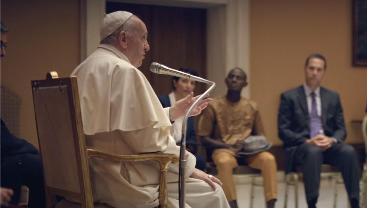 A imagem mostra o Papa Francisco em foco, com pessoas sentadas em sua volta, enquanto fala num microfone.