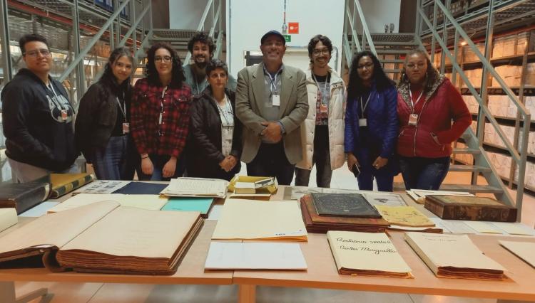 Alunos e professores posam para a foto no Arquivo Público do Estado de São Paulo. À frente deles, uma mesa com diversos livros e manuscritos.