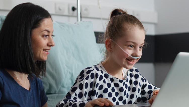Uma mulher branca, de cabelo preto e liso, sorrindo ao lado de uma criança branca, loira, do olho claro, que está usando um computador e fazendo oxigenoterapia em um hospital.