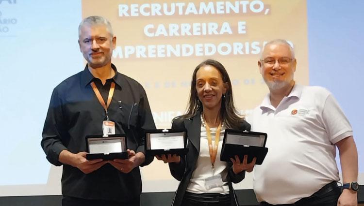 Professor Marcelo e Professora Edineide seguram as placas comemorativas ao lado de um representante da Associação Comercial