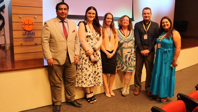 Sete pessoas posam para foto no auditório do UNIFAI, próximas ao púlpito com logo do centro universitário. Entre essas pessoas: professor Mário Jarrin, professora Karen Ambra, Rebeca, Professora Alessandra, Professor Alessandro e Professora Valéria.