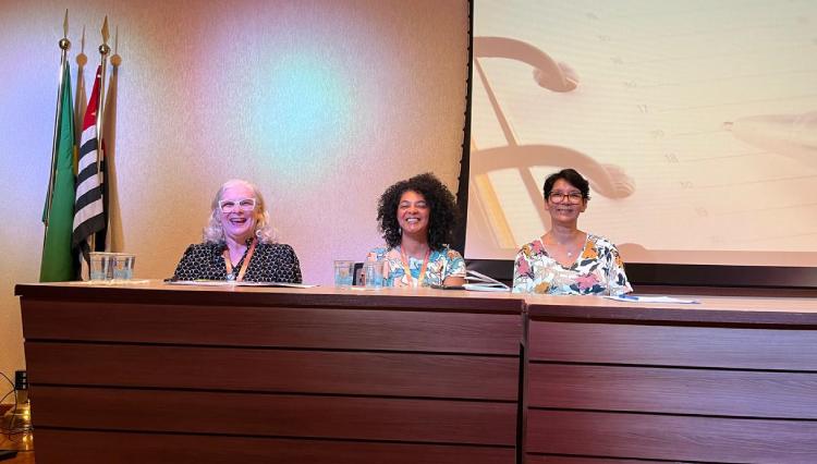 As três professoras palestrantes sentadas à mesa, no palco, posando para foto, antes do início das apresentações.